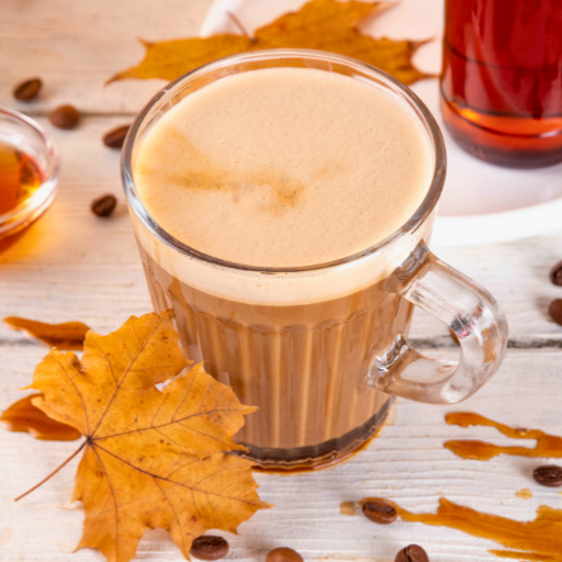 maple latte in a clear mug with froth on top and maple leaves and maple syrupon the table 