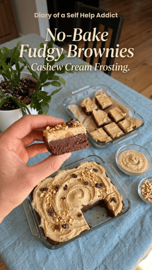 Close-up, appetizing food photography of a hand holding a single square of rich, dark chocolate brownie with one bite taken out of it. The brownie has a dense, fudgy texture and a thick, creamy white cashew frosting layer on top. In the softly blurred background, an 8x8-inch glass baking dish is filled with more brownies. Natural kitchen lighting, warm and inviting, realistic homemade food photography. Add elegant text overlay: “No-Bake Fudgy Brownies” with smaller text “+ Cashew Cream Frosting.”