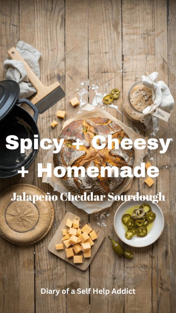 A beautiful overhead kitchen scene featuring the finished jalapeño cheddar sourdough alongside a Dutch oven, sourdough starter jar, banneton basket, parchment paper, cheddar cubes, and drained jalapeño peppers. Warm, rustic, approachable homemade-bread aesthetic.