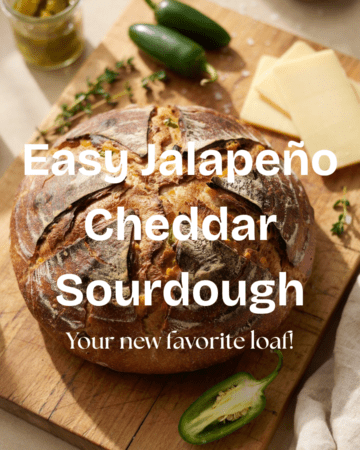 A rustic, golden-brown round loaf of jalapeño cheddar sourdough sitting on a wooden cutting board, with a few slices of creamy cheddar and fresh/pickled jalapeño peppers scattered around it. The crust is deeply browned and artisan-looking, with a beautiful scored design across the top. Warm, natural kitchen lighting and a cozy homemade-baking aesthetic.